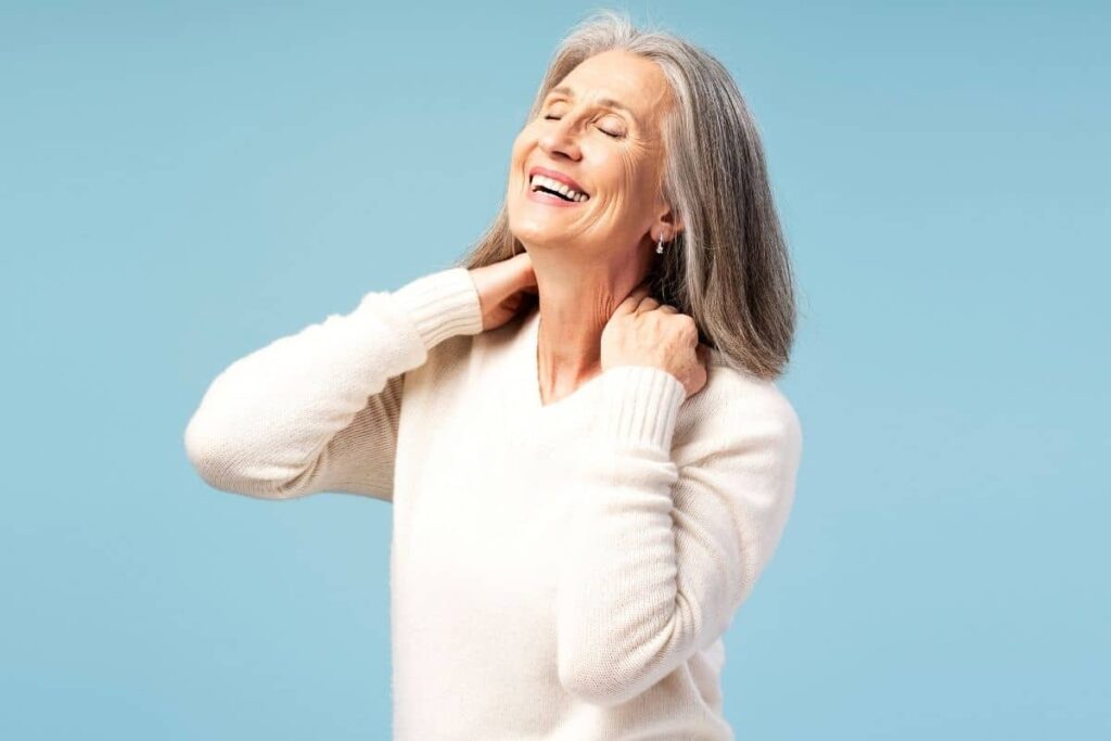 Mature woman smiling and touching her neck, showing confidence and comfort after neck lift recovery.