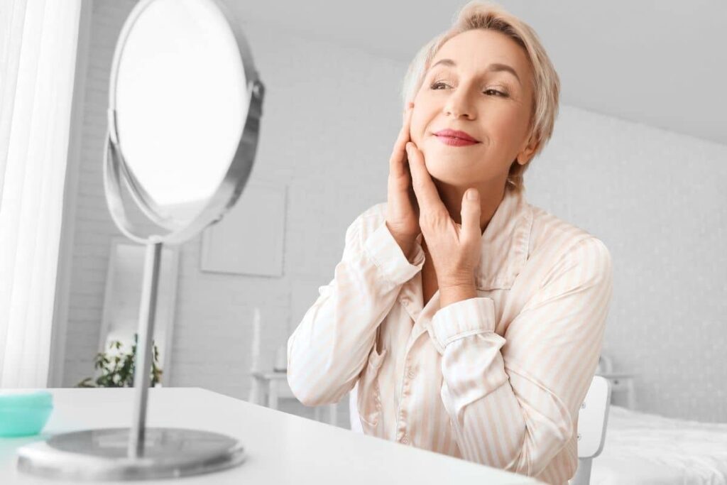 Confident older woman examining her face in mirror, pleased with combined neck lift and facial procedure results.