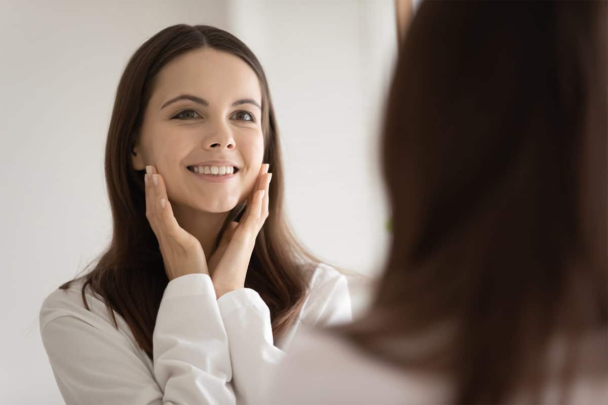 Happy young woman looking in a mirror touching her face after a Botox treatment.