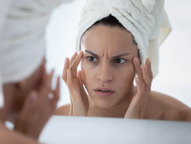 Woman with a towel on her head examining glabella wrinkles in the mirror, concerned about fine lines.