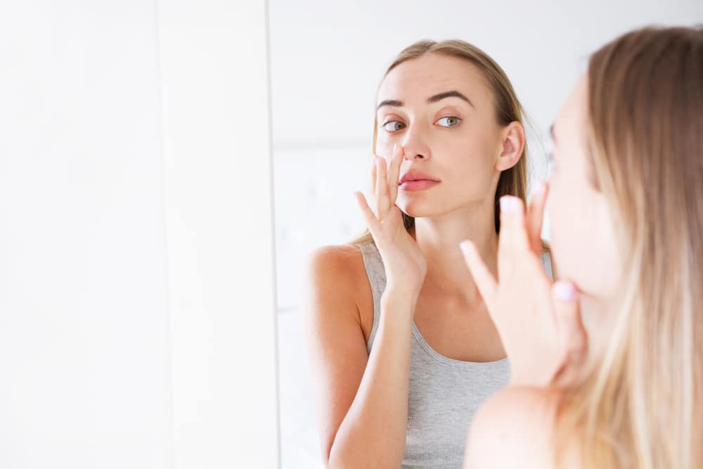 Woman examining her nose in the mirror while preparing for revision rhinoplasty with realistic expectations.