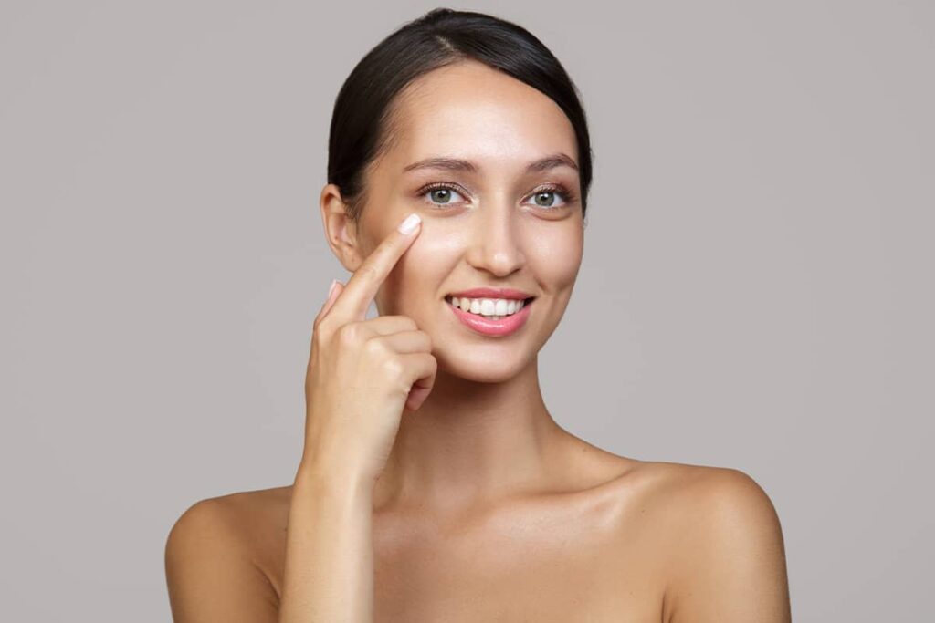 A smiling young woman looking at the camera and pointing to her lower eyelid area against a grey background.